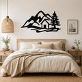 Black metal wall art silhouette of a mountain cabin scene with pine trees, mounted above a bed in a bedroom.