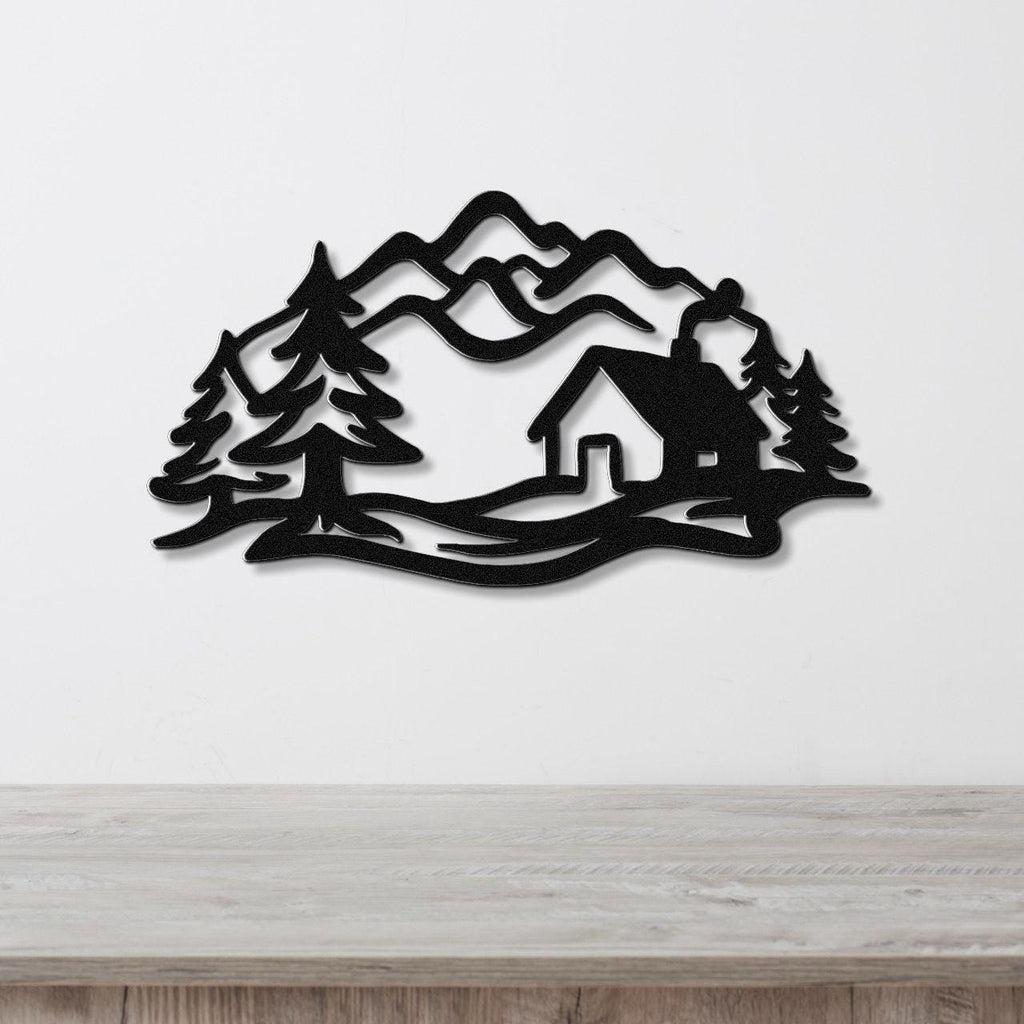 Black metal wall art featuring a mountain cabin silhouette with pine trees and layered peaks, mounted on a white wall above a wooden surface.