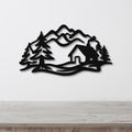 Black metal wall art featuring a mountain cabin silhouette with pine trees and layered peaks, mounted on a white wall above a wooden surface.