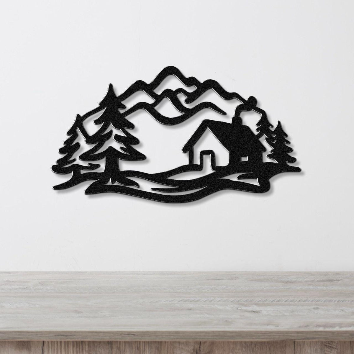 Black metal wall art featuring a mountain cabin silhouette with pine trees and layered peaks, mounted on a white wall above a wooden surface.