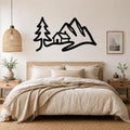 Black metal wall art silhouette of a mountain range with a cabin and pine tree, mounted above a bed in a neutral bedroom.