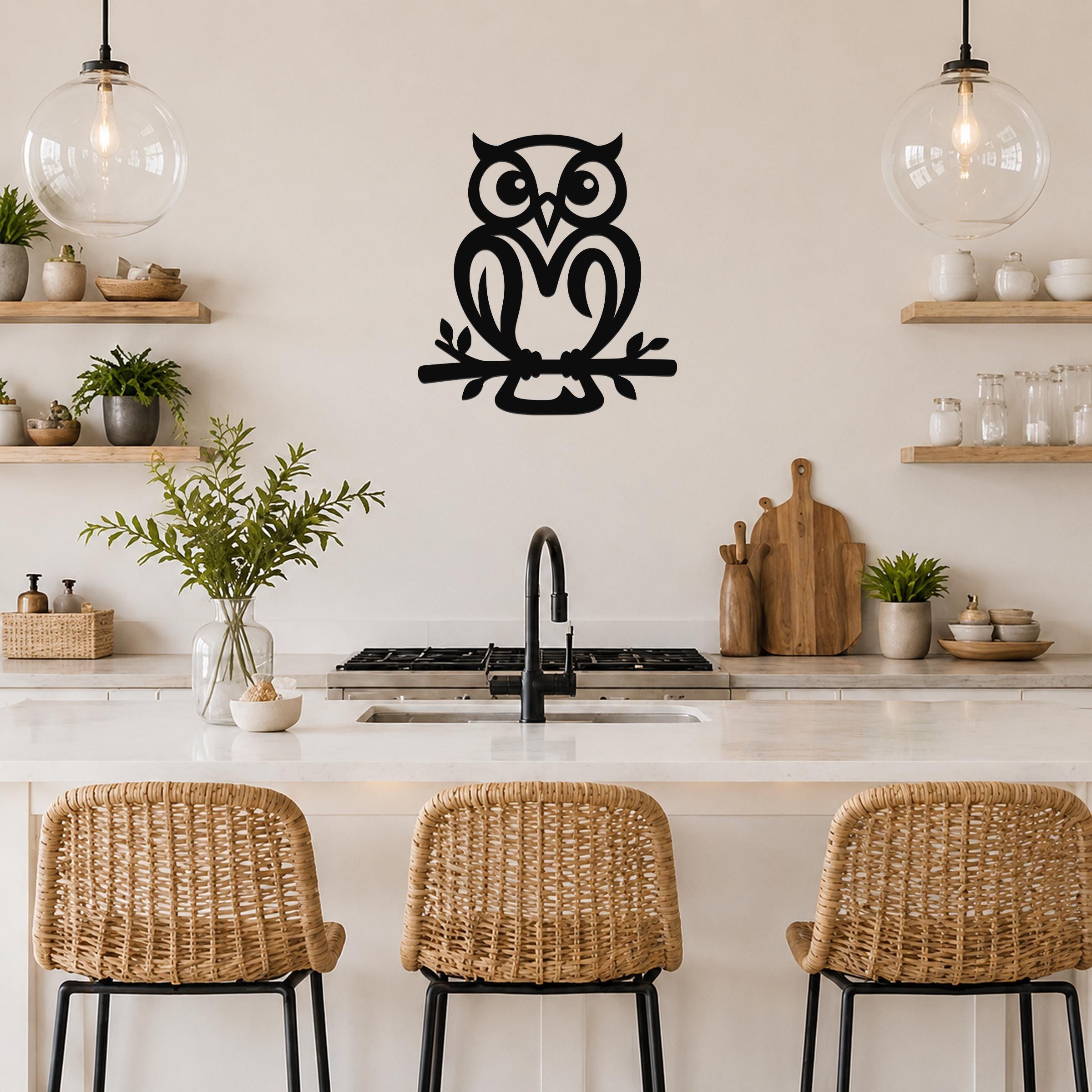 Black metal owl wall art with branch cutout, mounted on a kitchen wall above a counter with pendant lights and shelves.