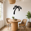 Black metal palm tree wall art silhouette mounted on a white wall above a round wooden table in a bright dining nook.