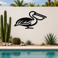 Black metal pelican wall art with a silhouette cutout design, mounted on a white outdoor wall beside a pool and desert plants.