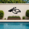 Black metal pelican wall art silhouette mounted on an outdoor wall above a poolside planter with purple flowers.