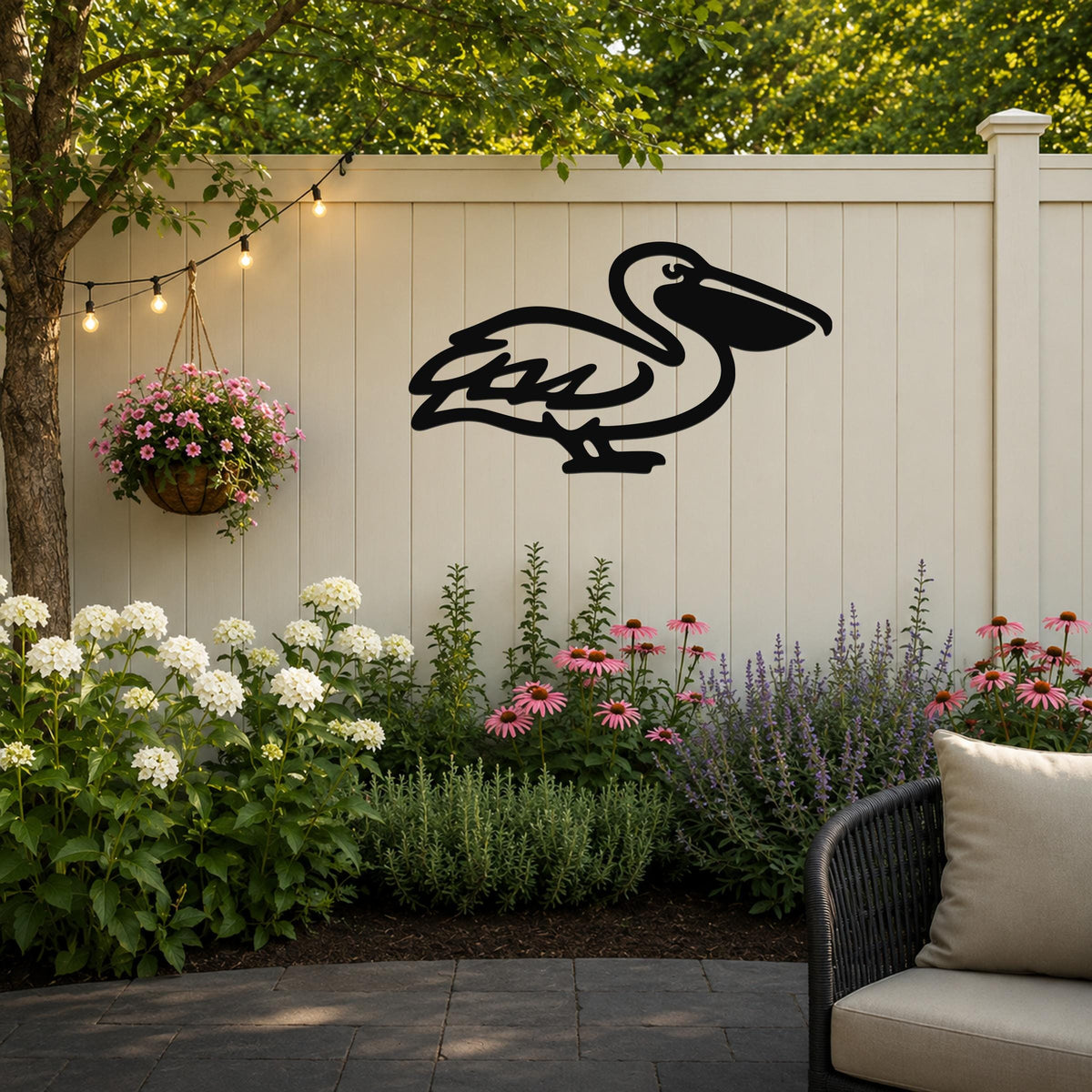 Black metal pelican wall art with a cutout silhouette, mounted on a light-colored outdoor fence above a garden bed with flowers and string lights.