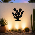 Black metal prickly pear cactus wall art with cutout details, mounted on a white outdoor wall above a desert garden with potted plants and ground lights.