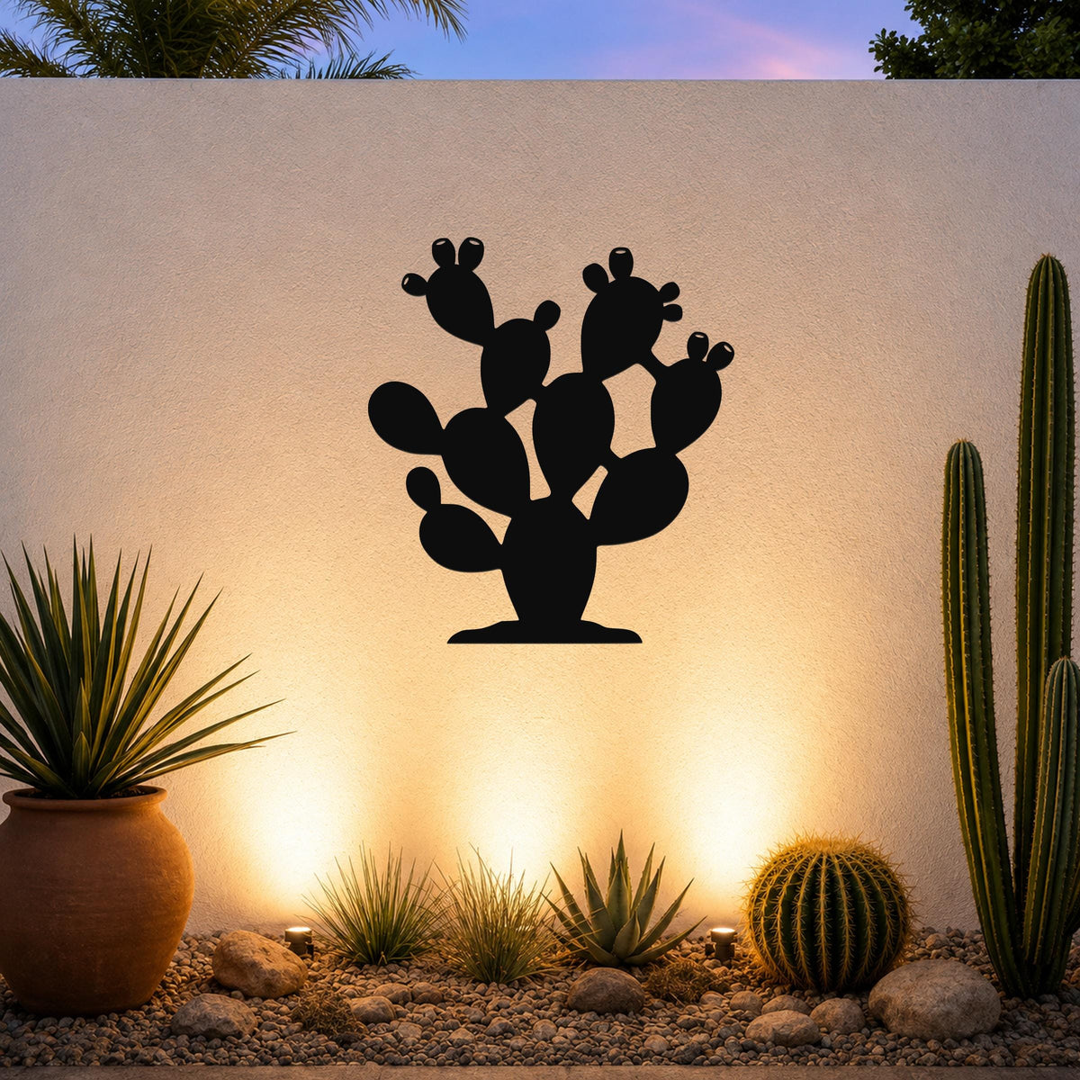 Black metal prickly pear cactus wall art with cutout details, mounted on a white outdoor wall above a desert garden with potted plants and ground lights.