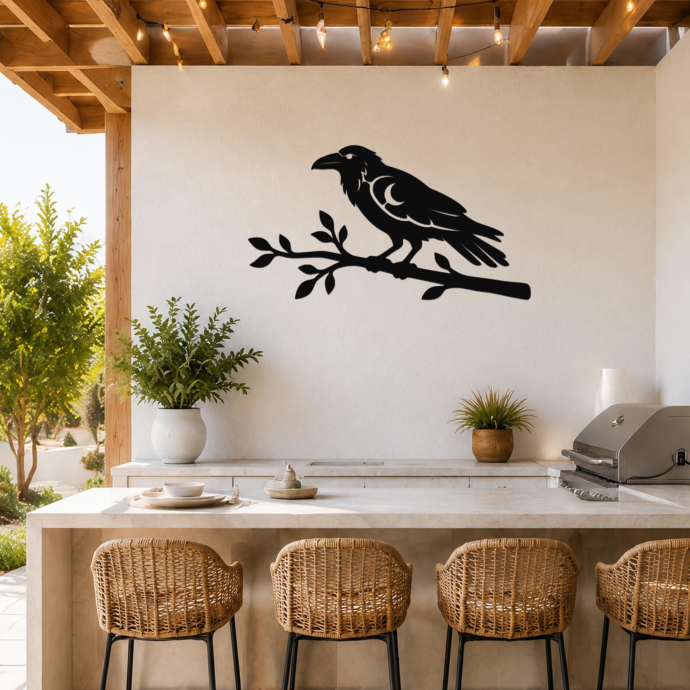Black metal raven wall art silhouette perched on a branch, mounted on a white outdoor kitchen wall above a counter with wicker stools.