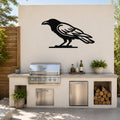 Black metal raven wall art silhouette mounted on an outdoor kitchen wall above a stainless steel grill and counter.