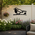 Black metal robin wall art silhouette perched on a branch, mounted on a light-colored outdoor fence above a garden bed with flowers and string lights.
