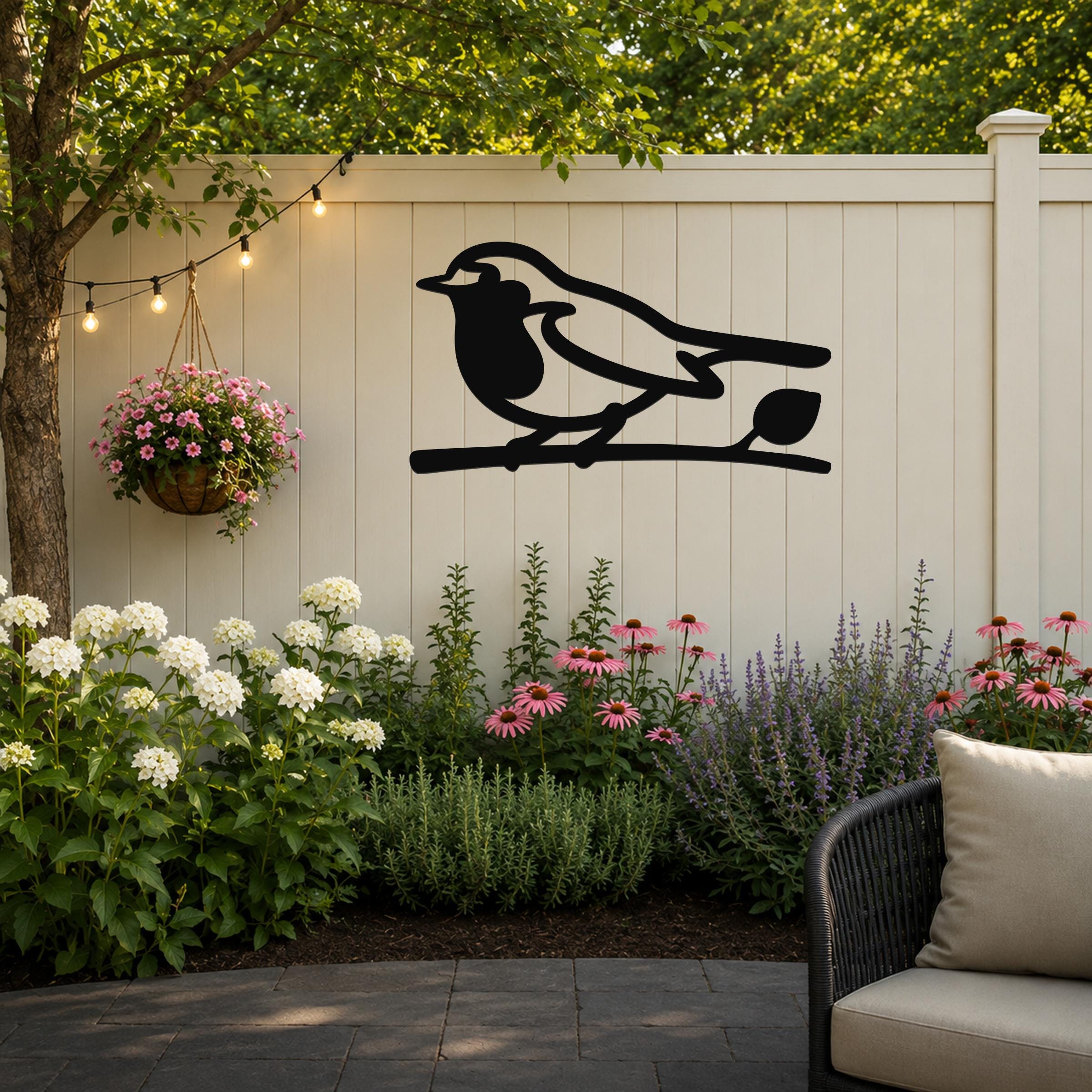 Black metal robin wall art silhouette perched on a branch, mounted on a light-colored outdoor fence above a garden bed with flowers and string lights.
