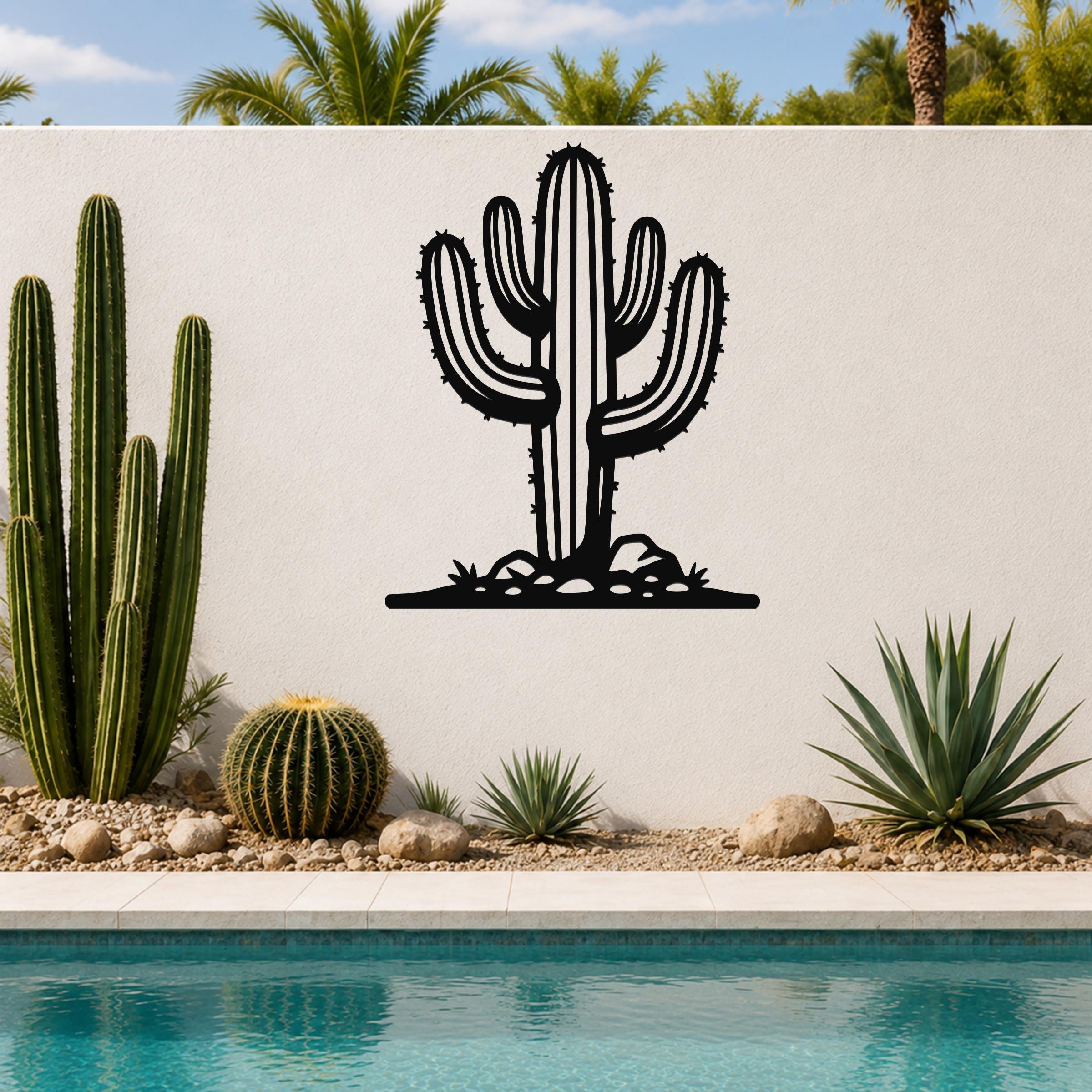 Black metal wall art in the silhouette of a saguaro cactus with branching arms and spines, mounted on a white outdoor wall above a poolside garden.