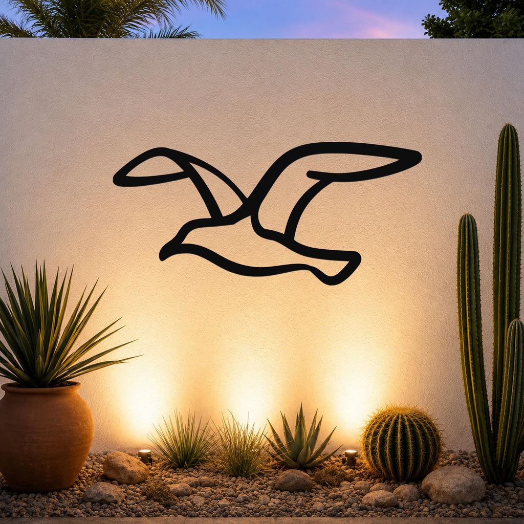 Black metal seagull wall art with a silhouette cutout design, mounted on an outdoor wall above a desert garden with cacti and warm uplighting.