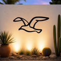 Black metal seagull wall art with a silhouette cutout design, mounted on an outdoor wall above a desert garden with cacti and warm uplighting.