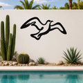 Black metal seagull wall art with flowing silhouette cutouts, mounted on an outdoor wall above a pool with cacti and palm trees in the background.