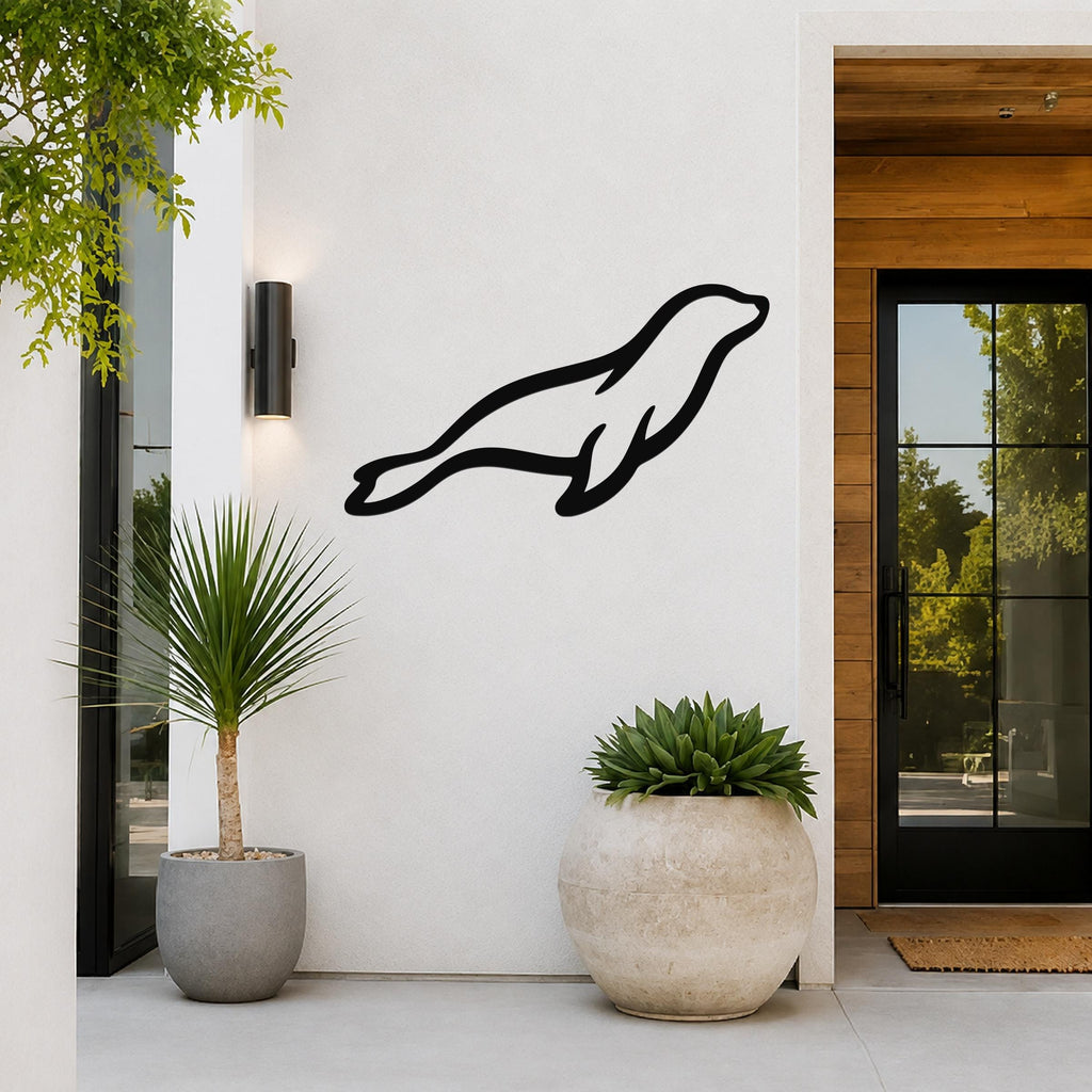 Black metal sea lion wall art silhouette mounted on a white exterior wall beside a glass door and potted plants.