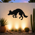 Black metal coyote wall art silhouette mounted on a textured outdoor wall above a desert garden with cacti and warm uplighting.