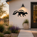 Black metal skunk silhouette wall art mounted on a white horizontal siding wall beside a front door, illuminated by a wall sconce at dusk.