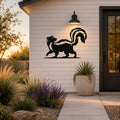 Black metal skunk silhouette wall art with a raised tail, mounted on a white siding exterior wall above a patio with plants and a pathway.