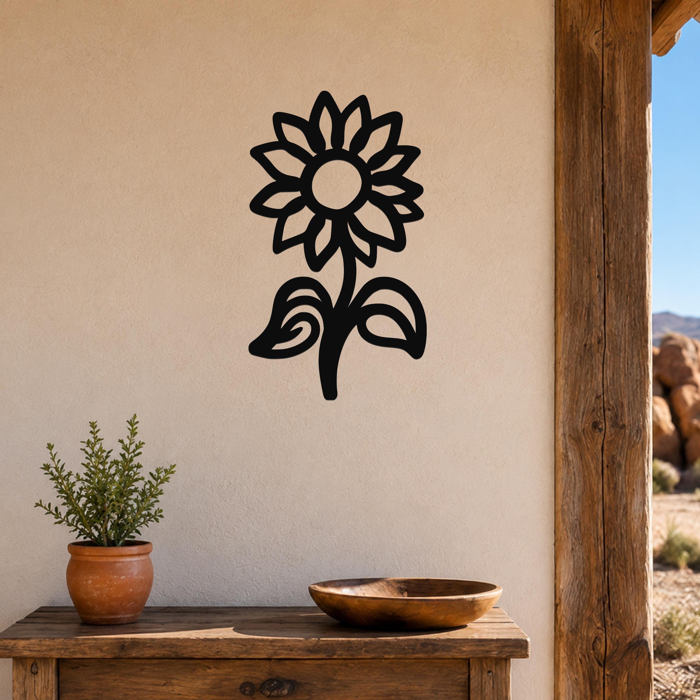 Black metal sunflower wall art with a silhouette design, mounted on a beige wall above a wooden table with a potted plant and bowl.