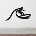 Black metal wall art cutout of a surfer riding a wave, mounted on a white wall above a light wood counter.