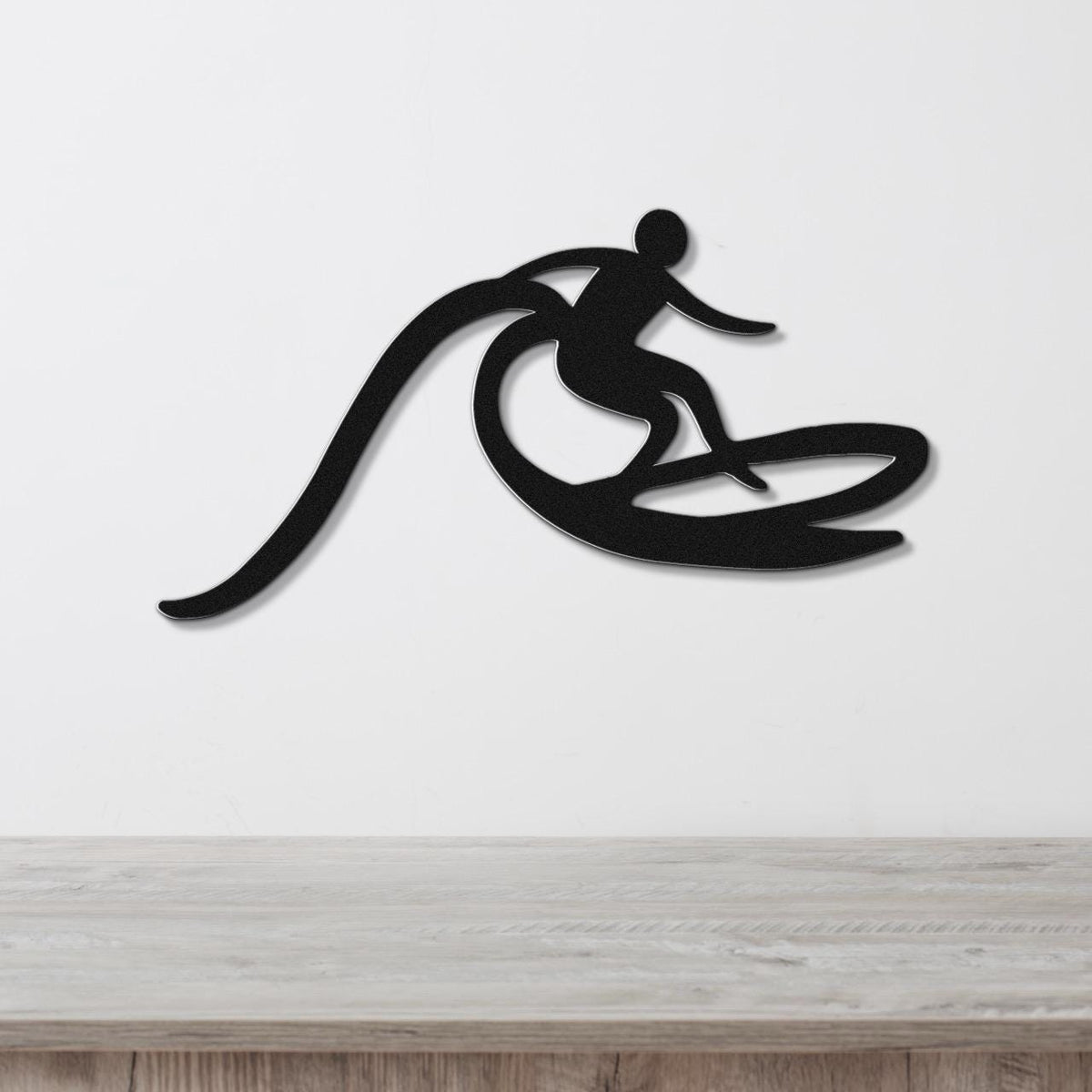 Black metal wall art cutout of a surfer riding a wave, mounted on a white wall above a light wood counter.