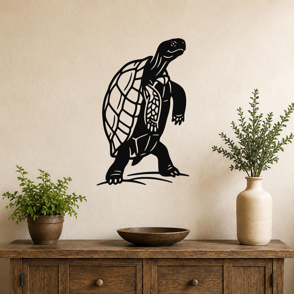 Black metal tortoise wall art with detailed shell and limb cutouts, mounted on a beige wall above a wooden console table with potted plants.