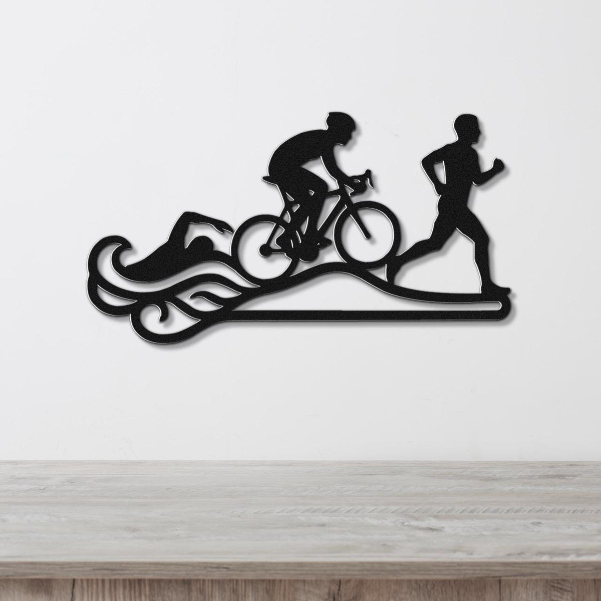 Black metal triathlon wall art with swim, bike, and run silhouettes and motion lines, mounted on a white wall above a wooden surface.