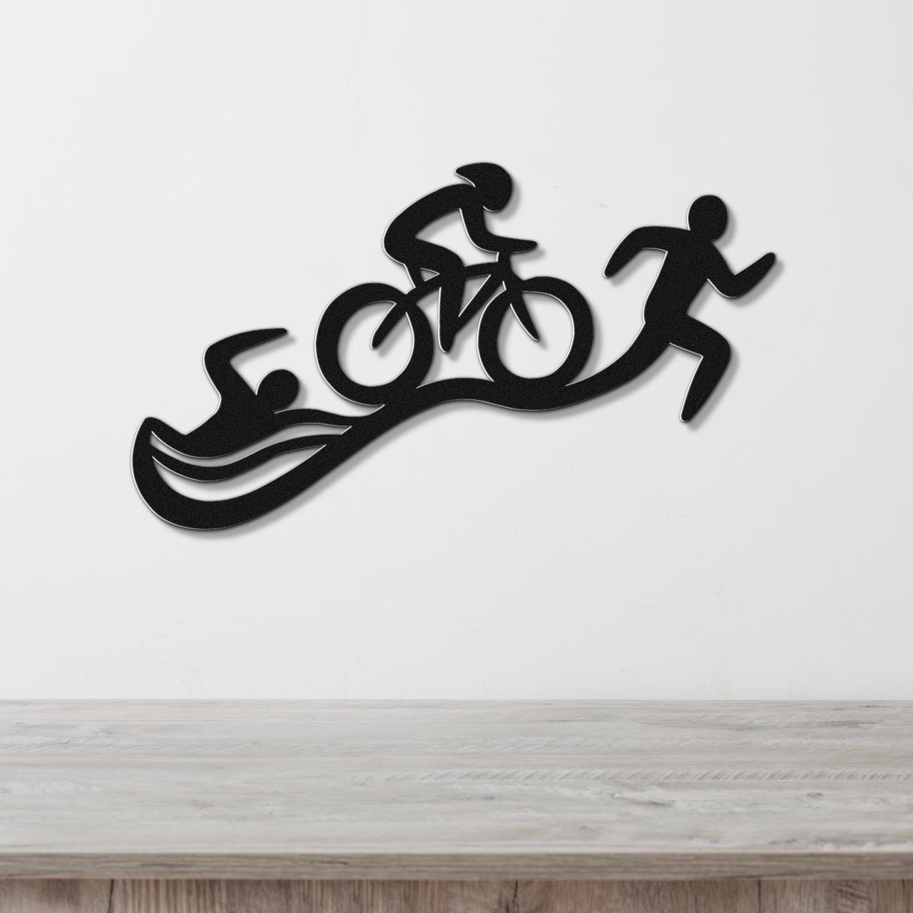 Black metal triathlon wall art with swim, bike, and run silhouettes, mounted on a white wall above a light wood surface.