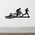Black metal wall art featuring a triathlon silhouette of swim, bike, and run figures with wave cutouts, mounted on a white wall above a wooden counter.