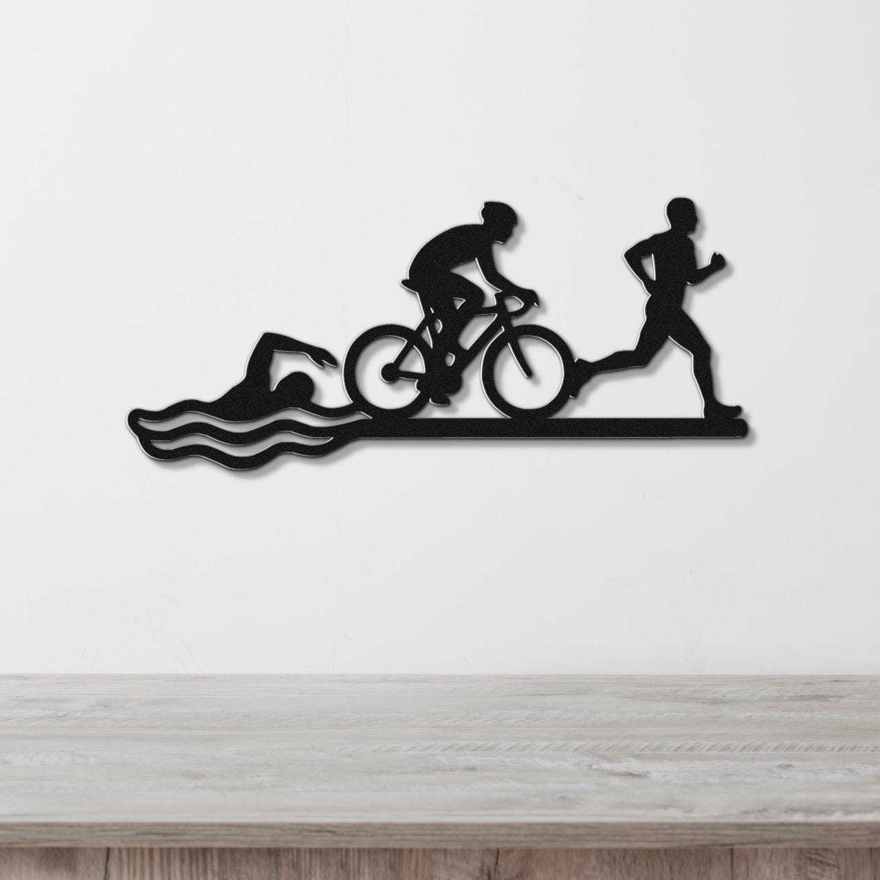 Black metal wall art featuring a triathlon silhouette of swim, bike, and run figures with wave cutouts, mounted on a white wall above a wooden counter.