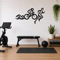 Black metal triathlon wall art with swim, bike, and run silhouette cutouts, mounted on a white shiplap wall above a gym bench in a home fitness room.