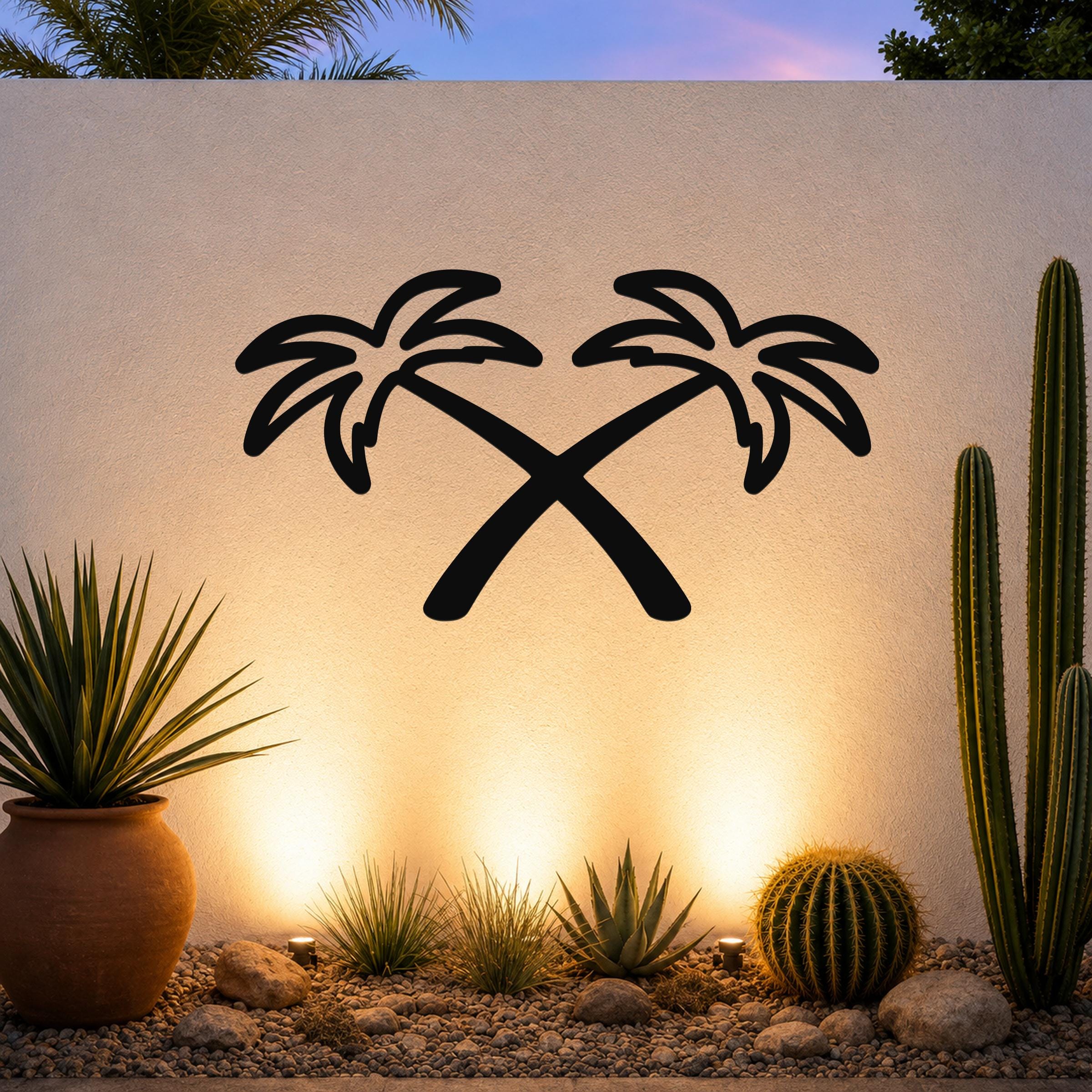 Black metal wall art featuring a silhouette of two crossed palm trees, mounted on an outdoor wall above a desert garden with cacti and warm uplighting.
