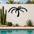Black metal palm tree wall art with curved frond cutouts, mounted on a white outdoor wall above a pool with cacti and succulents.