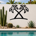 Black metal palm tree wall art with two crossed silhouettes, mounted on a white outdoor wall beside a pool and desert plants.