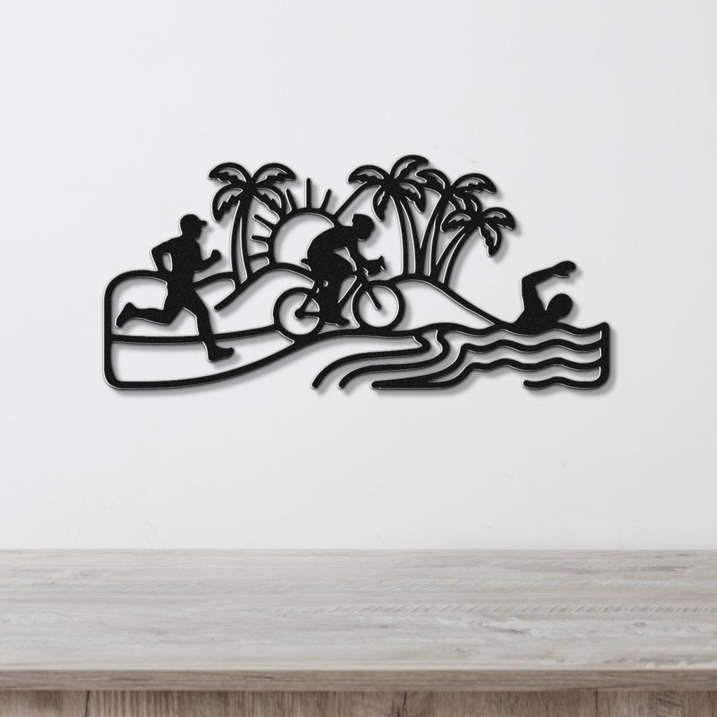 Black metal wall art cutout featuring a triathlon scene with swim, bike, and run silhouettes, palm trees, and waves, mounted on a white wall above a wooden surface.