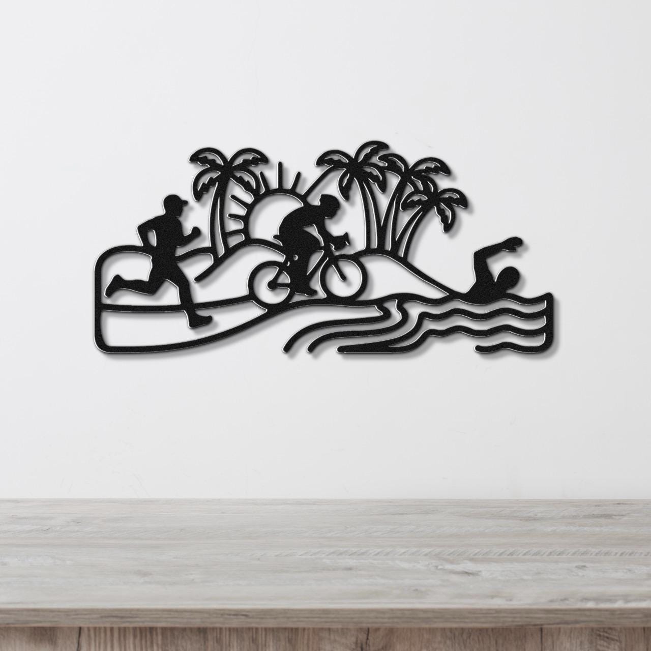 Black metal wall art cutout featuring a triathlon scene with swim, bike, and run silhouettes, palm trees, and waves, mounted on a white wall above a wooden surface.