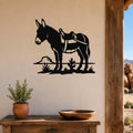 Black metal wall art silhouette of a burro with a saddle, cactus, and rock, mounted on a beige wall above a wooden table in a rustic indoor setting.