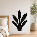 Black metal wall art in a silhouette of a snake plant, mounted on a white bedroom wall above a bed with a woven headboard.