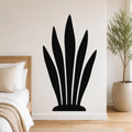 Black metal wall art in a silhouette of a snake plant, mounted on a white bedroom wall above a bed with a woven headboard.