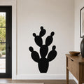 Black metal prickly pear cactus wall art with multiple paddle-shaped segments, mounted on a white wall in a living room near a door and console table.