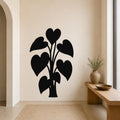 Black metal wall art in the silhouette of a heart-leaf philodendron plant, mounted on a beige living room wall above a woven rug.