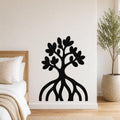 Black metal wall art in a tree silhouette with visible root and leaf cutouts, mounted on a white wall above a bed in a bedroom.