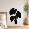 Black metal wall art in the silhouette of an elephant ear plant, mounted on a white wall above a bed in a bedroom.