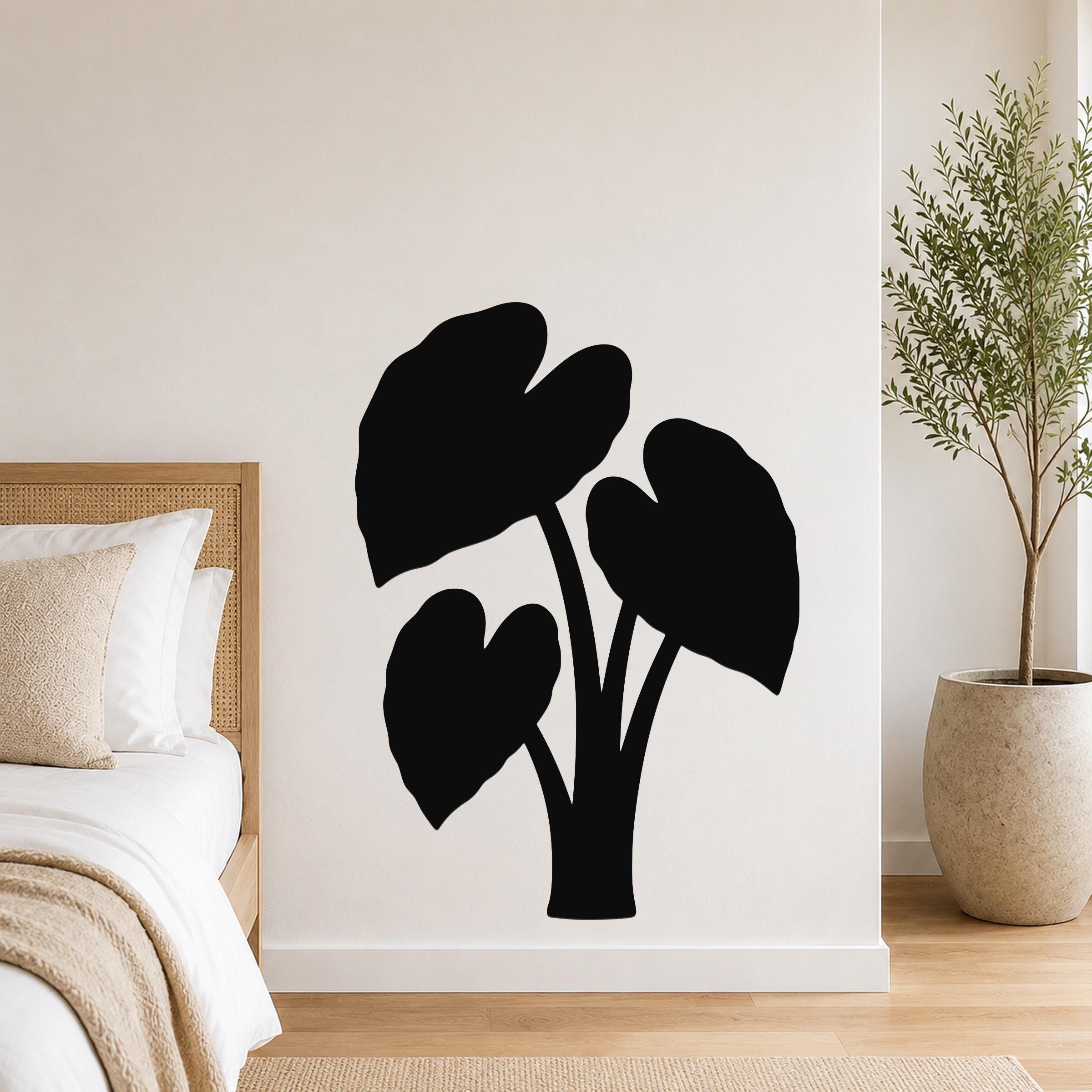 Black metal wall art in the silhouette of an elephant ear plant, mounted on a white wall above a bed in a bedroom.