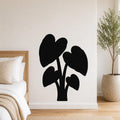 Black metal wall art silhouette of an elephant ear plant with large heart-shaped leaves, mounted on a white wall above a bed in a bedroom.