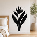 Black metal wall art in the silhouette of a bird of paradise plant, mounted on a white bedroom wall above a bed with a woven headboard.
