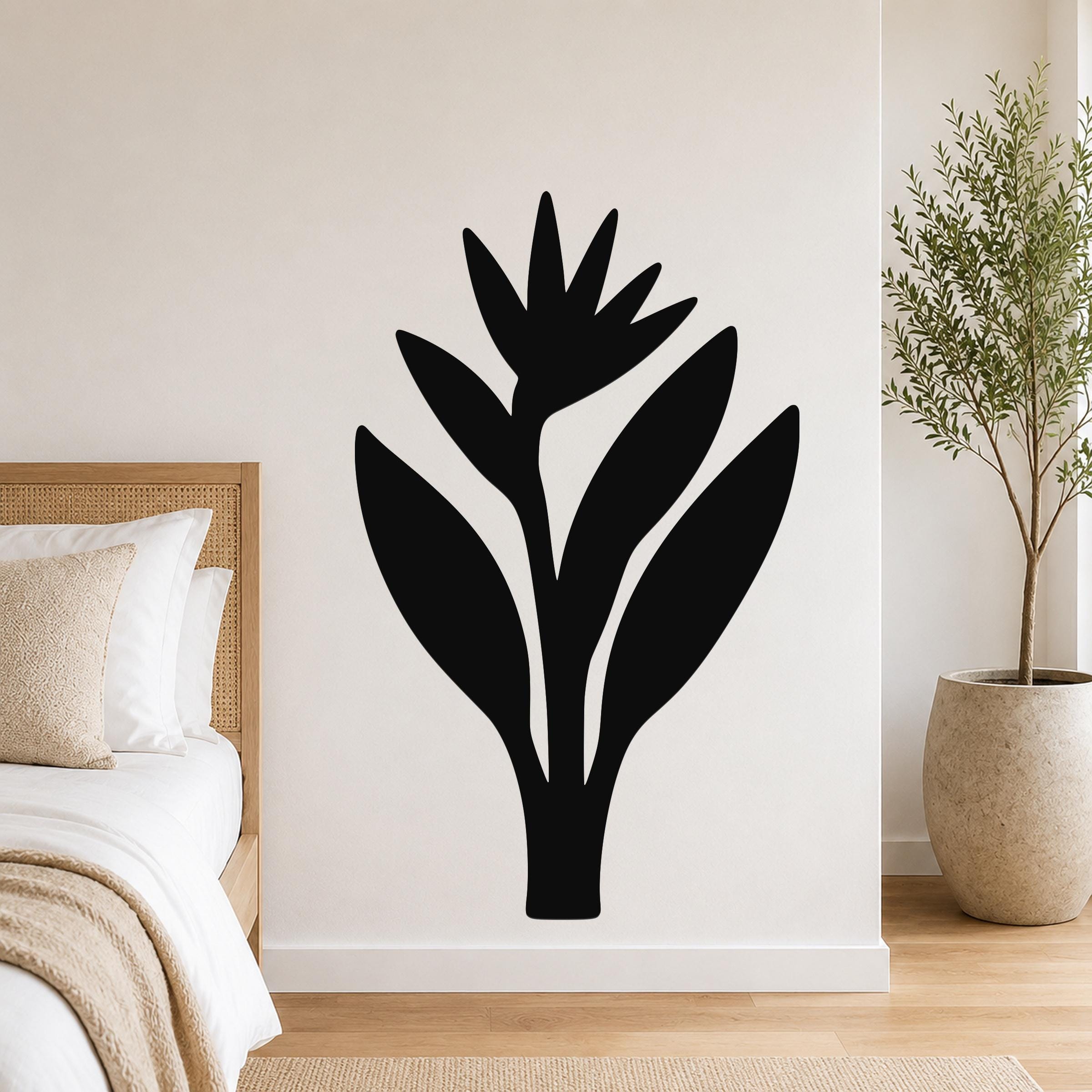 Black metal wall art in the silhouette of a bird of paradise plant, mounted on a white bedroom wall above a bed with a woven headboard.
