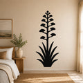 Black metal wall art in the silhouette of a blooming agave plant with a tall flower stalk and spiky leaves, mounted on a beige wall above a bed in a bedroom.